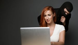 La mujer blanca pelirroja escribe en la computadora portátil mientras una figura amenazadora con pasamontañas mira sobre su hombro.